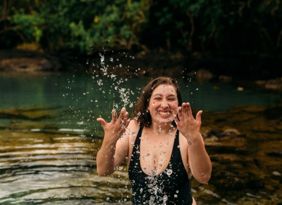 Eine junge plus size Frau steht im Badeanzug in einem See und spritzt Wasser hoch.