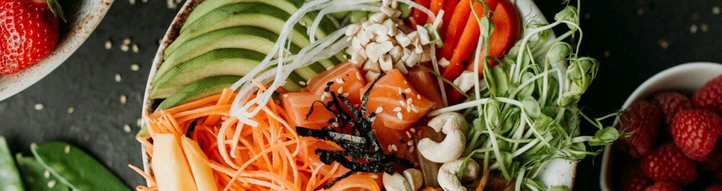 Bowl mit Avocado, Lachs und weiterem Gemüse auf einer dunklen Platte mit Obst