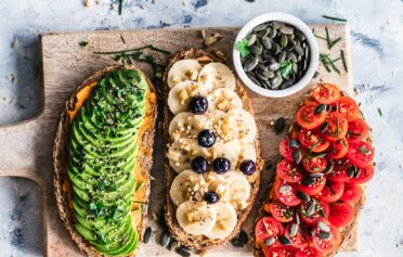 Drei Brote, eins mit Tomaten, eins mit Banane und eins mit Avocado belegt, liegen auf einem Holzbrett