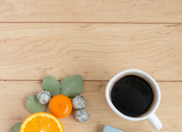 Auf einer Tischplatte liegt ein blaues Notizbuch, darauf steht eine Tasse Kaffe und ein Handy. Daneben liegt Obst.