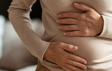 Frau mittleren Alters hält beide Hände am Bauch, sie hat die Augen geschlossen und einen Gesichtsausdruck von Unwohlsein