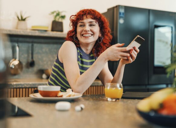 Junge rothaarige Frau hält am Essenstisch lachend ihr Smartphone in der Hand.