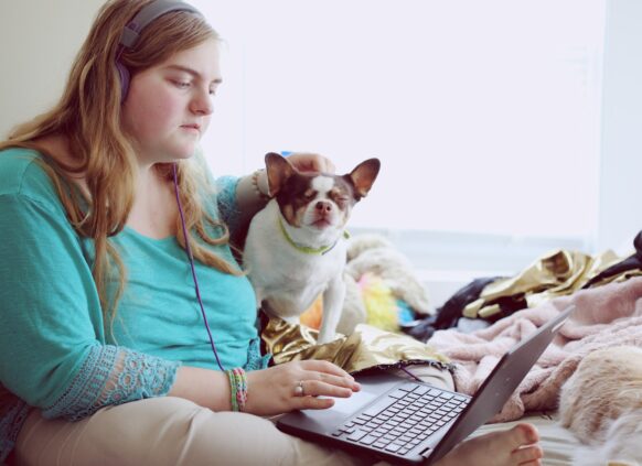 Übergewichtige Frau sitzt auf einem Sofa mit ihrem Hund und arbeitet am Laptop.