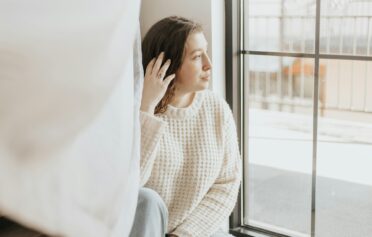 Eine junge Frau sitzt vor einem großen Fenster im Sonnenschein und sieht hinaus.