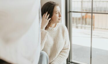 Eine junge Frau sitzt vor einem großen Fenster im Sonnenschein und sieht hinaus.