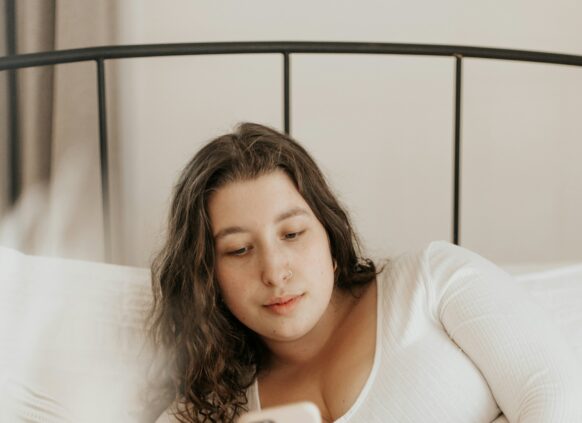 Eine junge plus size Frau liegt auf ihrem Bett und schaut auf ihr Handy.