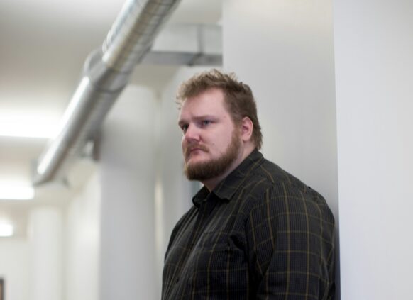 Ein plus size Mann lehnt an einer Wand und guckt in den Raum.