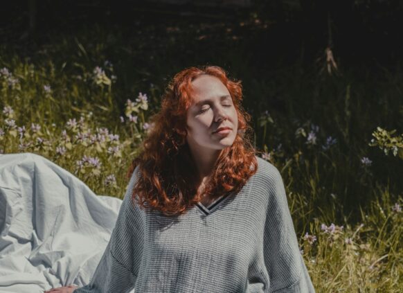 Eine junge Frau sitzt auf einer Picknick Decke in der Sonne und hält hr Gesicht genussvoll Richtung Sonne.