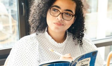 Eine junge Frau sitzt mit einem Buch vor einem Fenster und blickt nachdenklich zur Seite.