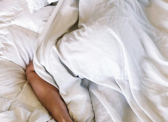 Eine Person liegt schlafend im Bett und ist bedeckt mit der Bettdecke, nur ein Bein guckt heraus.