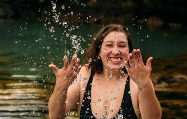 Eine junge plus size Frau in einem schwarzen Badeanzug steht in einem See und spritzt fröhlich lächelnd Wasser in die Luft.