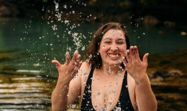 Eine junge plus size Frau in einem schwarzen Badeanzug steht in einem See und spritzt fröhlich lächelnd Wasser in die Luft.