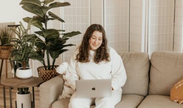 Eine junge plus size Frau sitzt auf ihrem Sofa mit ihrem Laptop auf dem Schoß und schaut lächelnd auf den Bildschirm.