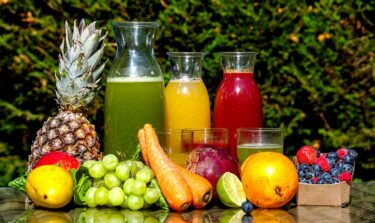 Frisches Obst und Gemüse und drei Karaffen mit frischem Saft und Gläsern sind auf einem Gartentisch drapiert.
