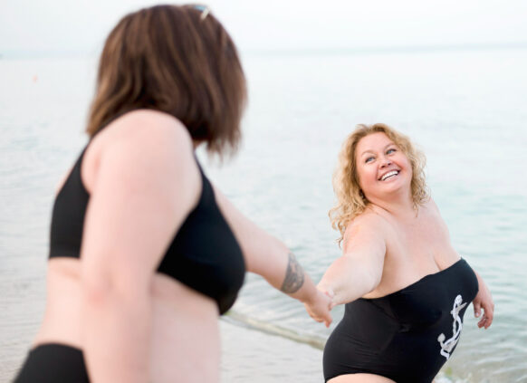 Eine plus size Frau steht im Badeanzug am Strand und hält eine andere Frau an der Hand.