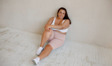 Eine junge plus size Frau sitzt in workout Kleidung auf dem Boden und lächelt.
