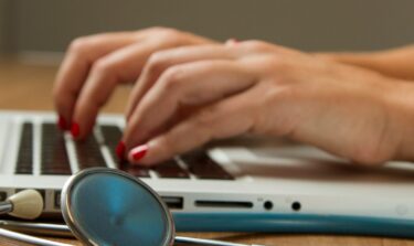 Eine Nahaufnahme von Händen, die auf einem Laptop tippen und daneben liegt ein Stetoskop.