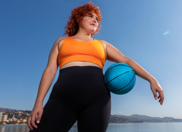 Eine kurvige Frau von unten abgelichtet steht vor einem See, der Himmel ist blau. Sie trägt Sportkleidung und hat unter ihrem linken arm einen Basketball klemmen.
