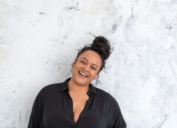 Eine junge curvy Frau steht lässig vor einer Wand und lächelt in die Kamera.