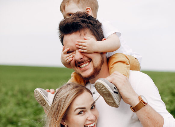 Ein Bild einer jungen Familie, auf dem die Frau ihren Mann umarmt, der auf den Schulter ihr Kind trägt. Alle lachen fröhlich.