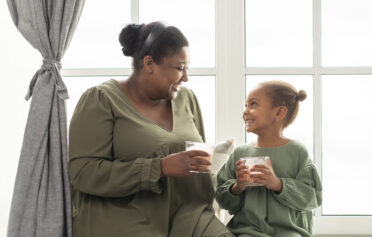 Eine Mutter sitzt neben ihrer Tochter auf einer Fensterbank. Beide halten ein Glas in der Hand und lachen sich fröhlich an.