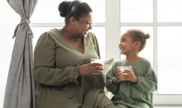 Eine Mutter sitzt neben ihrer Tochter auf einer Fensterbank. Beide halten ein Glas in der Hand und lachen sich fröhlich an.