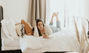 Junge Frau liegt auf dem Bett und schaut auf ihr Handy
