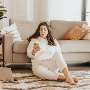 Frau sitzt mit Handy neben Couch und sucht Tipps um Bauchfett loszuwerden