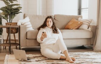 Frau sitzt mit Handy neben Couch und sucht Tipps um Bauchfett loszuwerden