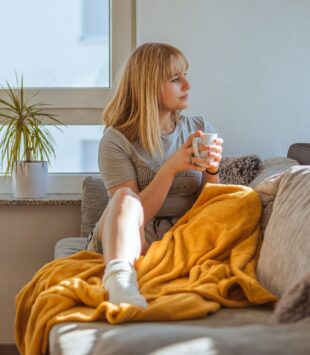 Frau sitzt auf einem Sofa und schaut zur Seite, über ihr liegt eine gelbe Decke.