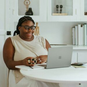 Eine Frau im Kleid sitzt an einem Tisch vor ihrem Laptop und lacht.