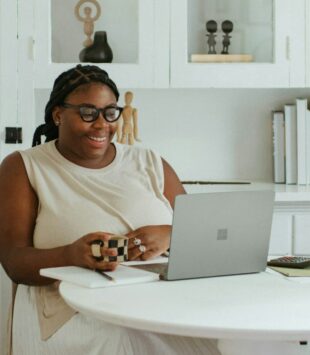 Eine Frau im Kleid sitzt an einem Tisch vor ihrem Laptop und lacht.