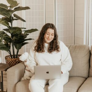 Eine junge plus size Frau sitzt auf ihrem Sofa mit ihrem Laptop auf dem Schoß und schaut lächelnd auf den Bildschirm.