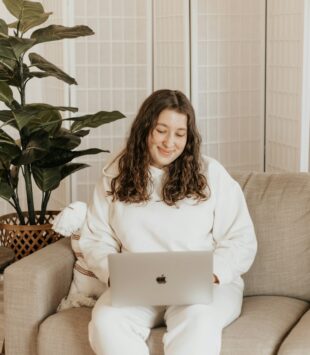 Eine junge plus size Frau sitzt auf ihrem Sofa mit ihrem Laptop auf dem Schoß und schaut lächelnd auf den Bildschirm.