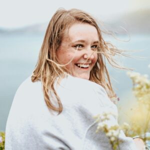 Frau sitzt auf einer Klippe an Strand und schaut lachend über die Schulter.