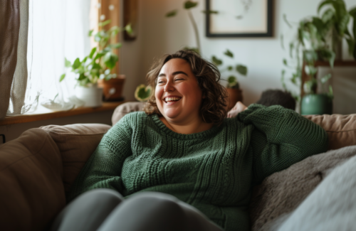 Adipositöse Frau sitzt in einem gemütlichen Wohnzimmer lachend auf einem Sofa