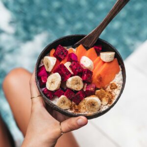 Eine Hand hält eine Schüssel mit vielen Früchten in der Hand, im Hintergrund ist ein Pool zu sehen.