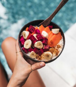 Eine Hand hält eine Schüssel mit vielen Früchten in der Hand, im Hintergrund ist ein Pool zu sehen.
