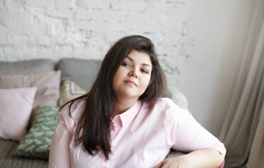 Eine junge Frau sitzt seitlich auf dem Sofa und schaut selbstbewusst in die Kamera.