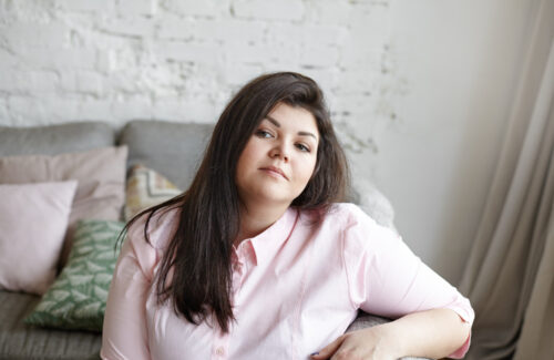 Eine junge Frau sitzt seitlich auf dem Sofa und schaut selbstbewusst in die Kamera.