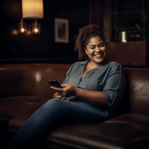 Eine poc plus size Frau sitzt auf einem Ledersofa und lächelt zur Seite, während sie ihr Handy in der Hand hält.