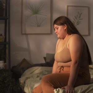 Eine plus size Frau sitzt in gemütlicher Kleidung auf einem Bett und schaut nach unten.