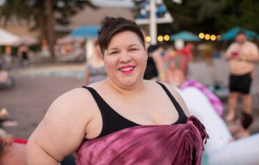 Eine selbstbewusste plsu size Frau trägt einen Badeanzug und ein umgewickeltes Handtuch. Im Hintergrund ist ein Schwimmbad zu erkennen. Sie lächelt fröhlich in die Kamera.