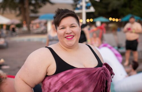 Eine selbstbewusste plsu size Frau trägt einen Badeanzug und ein umgewickeltes Handtuch. Im Hintergrund ist ein Schwimmbad zu erkennen. Sie lächelt fröhlich in die Kamera.