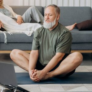 Ein älterer Herr meditiert im Schneidersitz auf einer Yogamatte im Wohnzimmer.