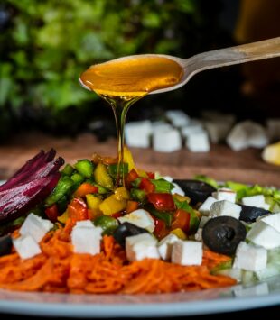 Ein meditteraner Salat mit Oliven auf dem ein Löffel Olivenöl verteilt wird.