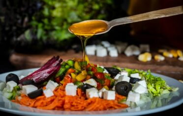 Ein meditteraner Salat mit Oliven auf dem ein Löffel Olivenöl verteilt wird.