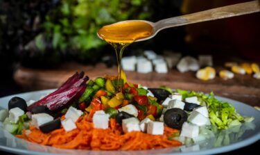 Ein meditteraner Salat mit Oliven auf dem ein Löffel Olivenöl verteilt wird.