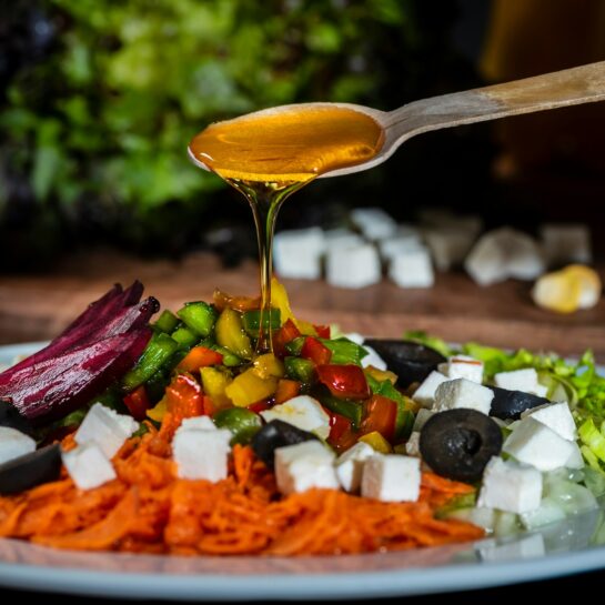 Ein meditteraner Salat mit Oliven auf dem ein Löffel Olivenöl verteilt wird.