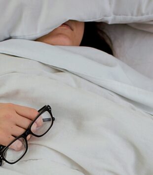 Frau liegt erschöpft im Bett mit Brille in der Hand